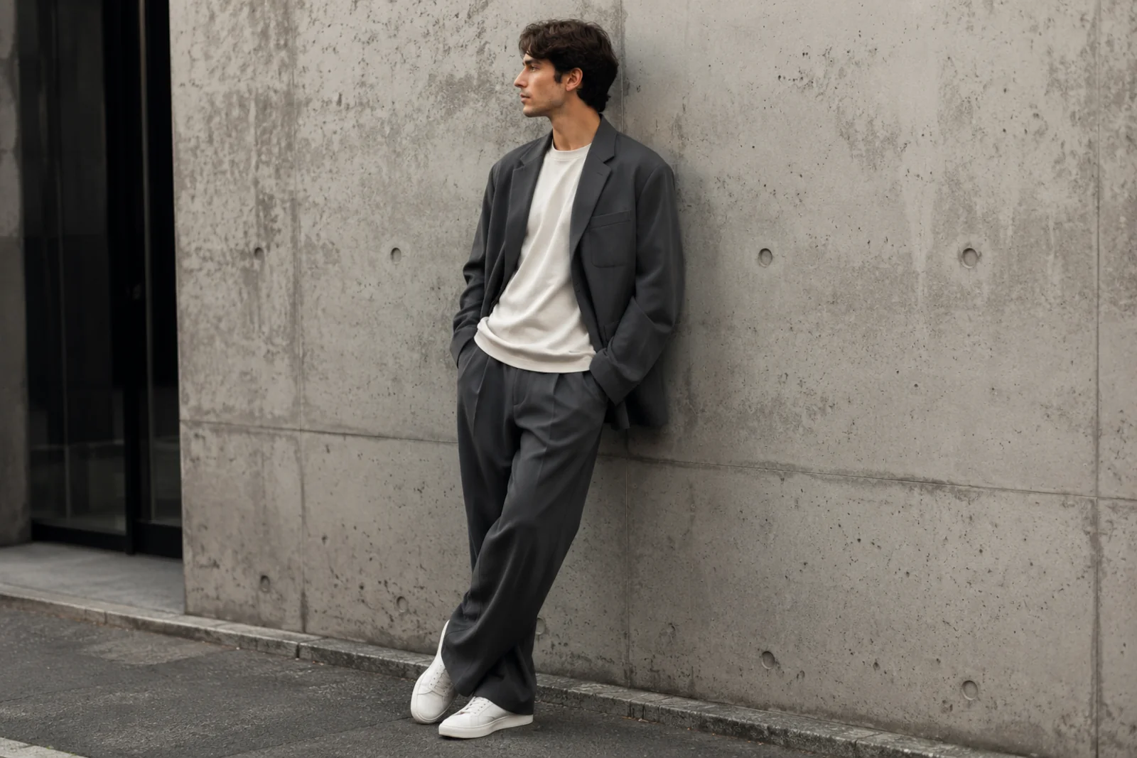 Mężczyzna w szarym garniturze oversize, białym T-shircie i sneakersach stojący przy betonowej ścianie